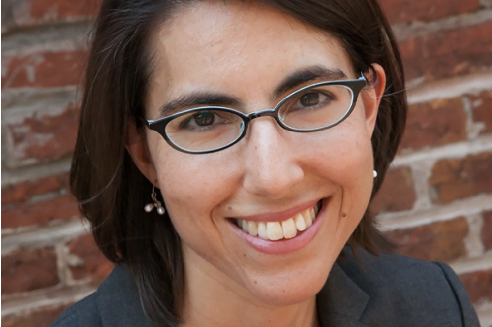 Close-up portrait of smiling woman with glasses against brick wall.